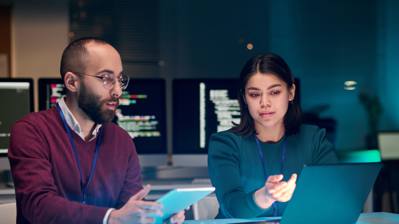 Two cybersecurity analysts working together with a tablet and laptop in a high-security environment.